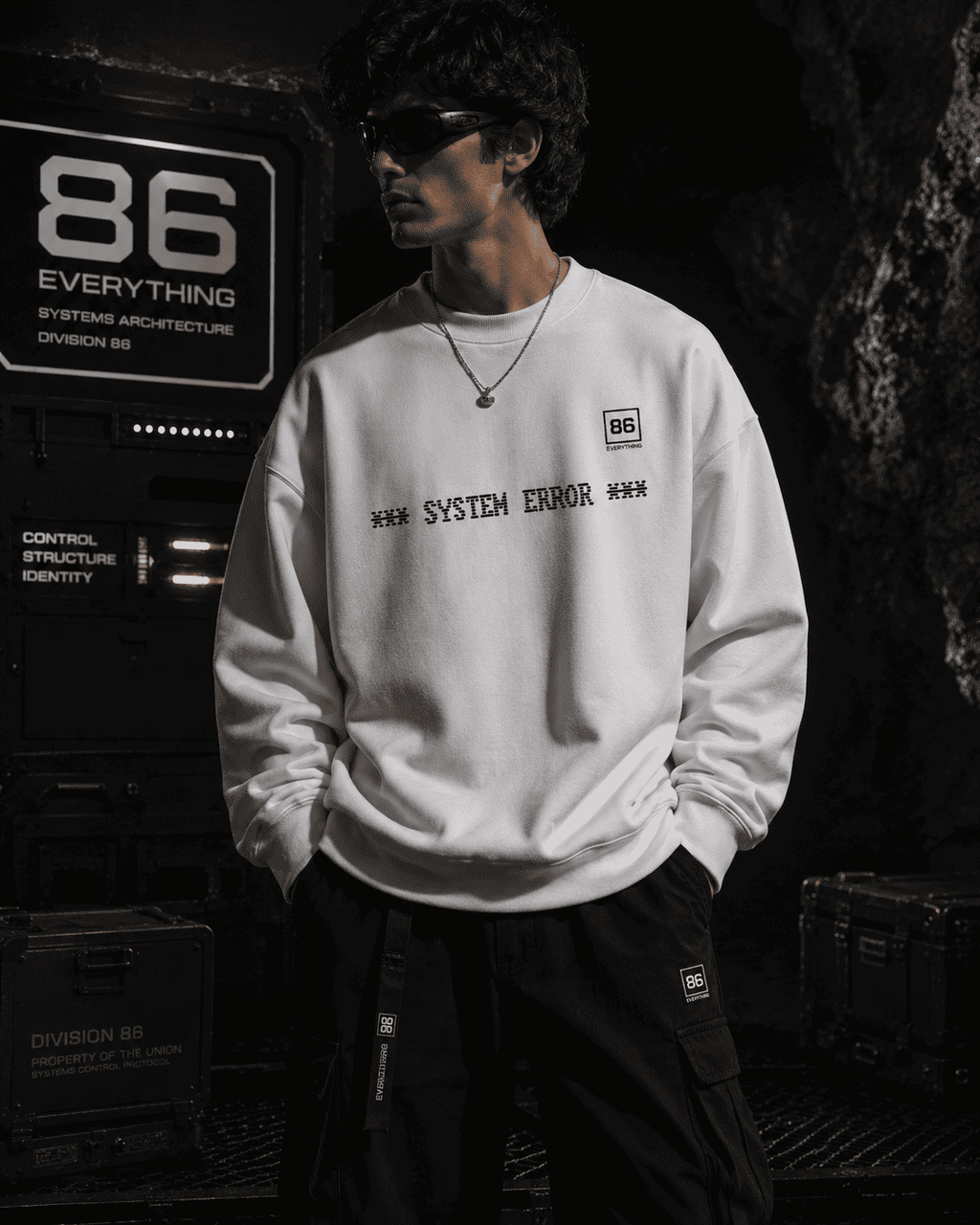 White fabric sweatshirt with SYSTEM ERROR printed on the chest, worn by a man in a dark industrial setting.
