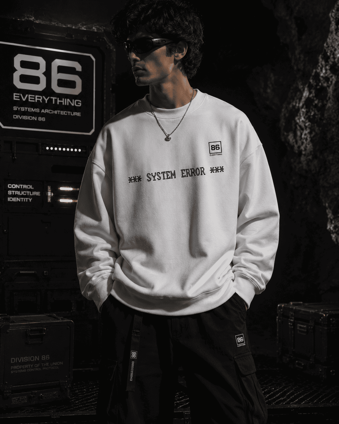 White fabric sweatshirt with SYSTEM ERROR printed on the chest, worn by a man in a dark industrial setting.